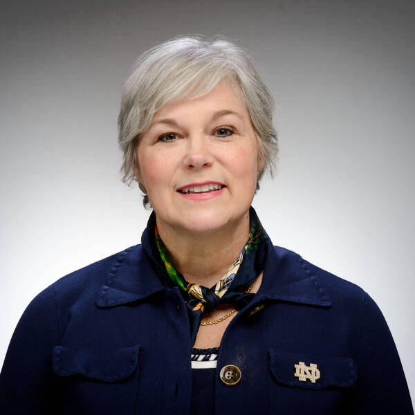 Smiling woman with short gray hair, wearing a navy blue top, a colorful scarf, and a gold Notre Dame ND pin.