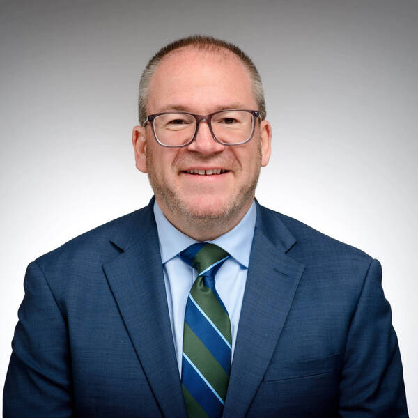 Smiling man with glasses, short beard, wearing a blue suit, light blue shirt, and blue/green striped tie.
