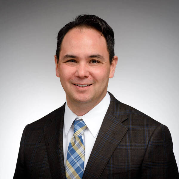 A smiling man with dark hair wears a brown plaid suit jacket, white shirt, and blue and yellow plaid tie.