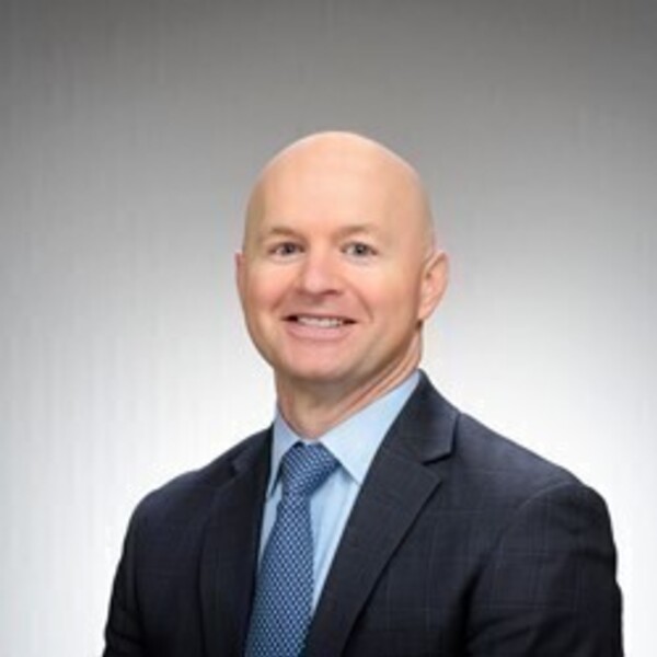 A bald man smiles, wearing a dark blue suit, light blue shirt, and patterned blue tie.
