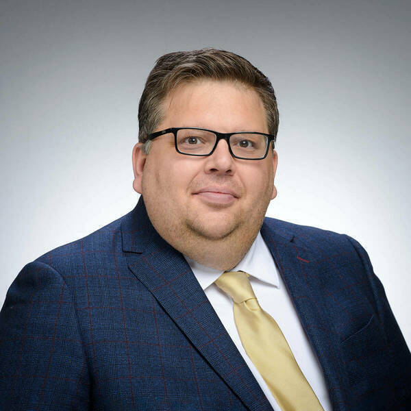 Smiling man with dark hair, black glasses, dark blue suit, white shirt, and gold tie on gray background.