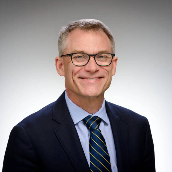 Professional man with grey hair, black glasses, dark blue suit, light blue shirt, and blue and gold striped tie, smiling.