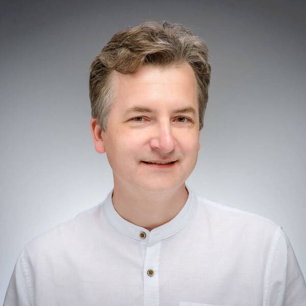 A smiling man with salt-and-pepper hair wears a white linen collared shirt with wooden buttons against a gray background.