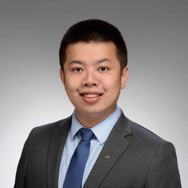 Headshot of Toby Li, A smiling man with short dark hair in a grey suit, blue checkered shirt, blue tie, and a gold Notre Dame pin on his lapel.
