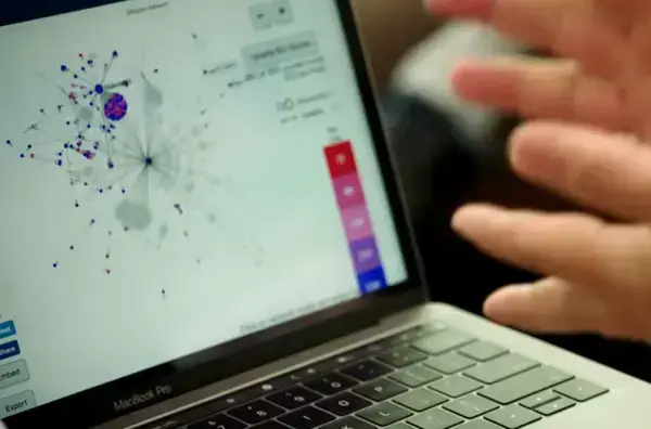 A laptop screen shows a network graph with blue and red nodes. A hand gestures over the keyboard.