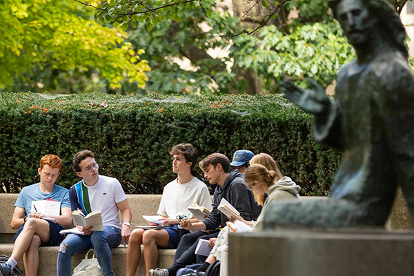 Students study books on a stone bench at Notre Dame, with trees, hedges, and a blurred Christ statue.