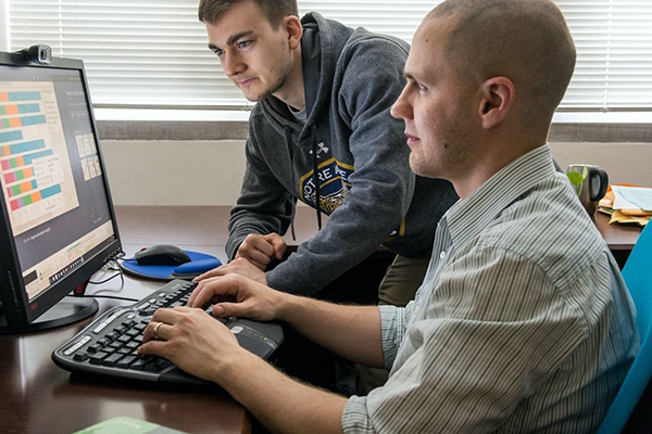 Two men intently look at a computer screen displaying colorful charts. One types while the other, in a Notre Dame hoodie, leans over.