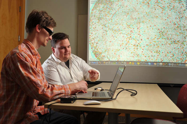 Man in orange plaid shirt and sunglasses works on laptop. Man in white shirt points at screen. Large colorful data visualization projected