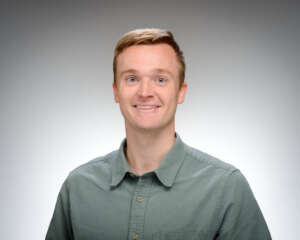 A smiling young man with light skin and short light brown hair wears a teal button-down shirt