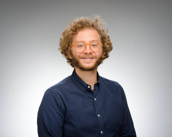 Headshot of Numo Moniz, a man with curly reddish-blonde hair, a beard, and orange glasses smiles, wearing a dark blue button-up shirt.