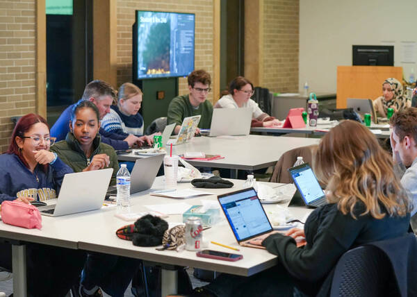 Notre Dame students collaborate and study on laptops at white tables. A smiling student in a blue ND jacket talks with another in a green jacket.
