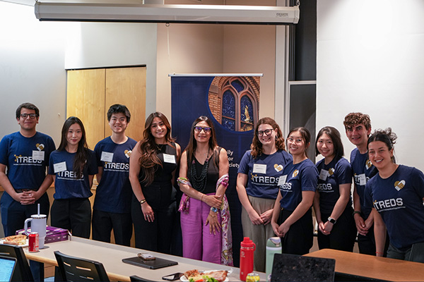 Nine smiling students in navy 'TREDS' shirts and a woman in pink pants stand before a Notre Dame Basilica banner.