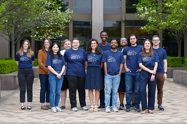 A diverse group of 12 people in blue LUCY shirts smile, standing outdoors by a modern building.