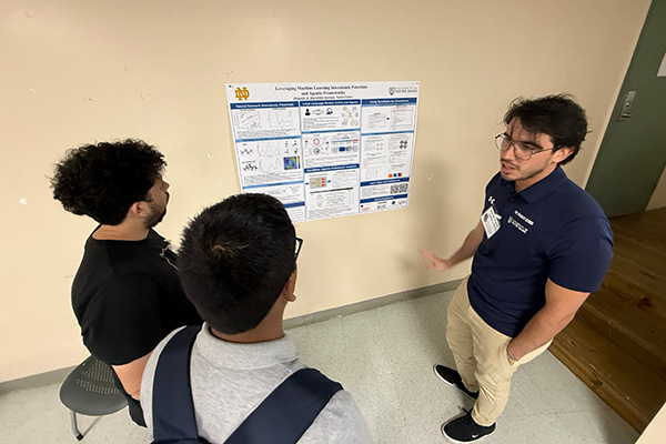 A man in a navy polo and glasses gestures while explaining a Notre Dame research poster to two people listening intently.