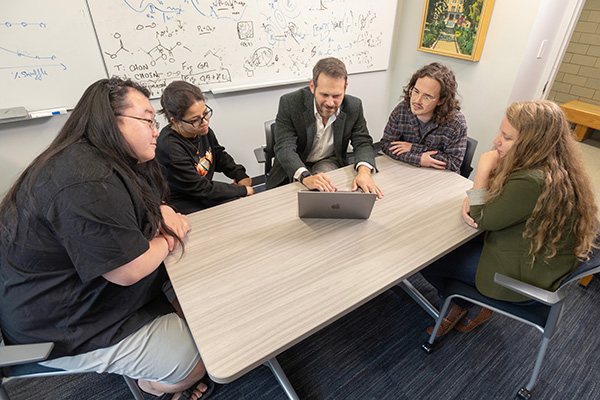 Professor in green blazer shows laptop to four attentive students gathered at a table with equations on a whiteboard.