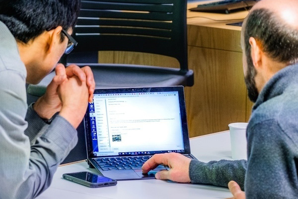 Two men look at a laptop screen. One in a grey shirt with glasses has clasped hands; the bearded man uses the touchpad.