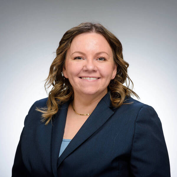 Smiling woman with wavy brown hair, wearing a dark blue blazer, light blue top, and a gold necklace.