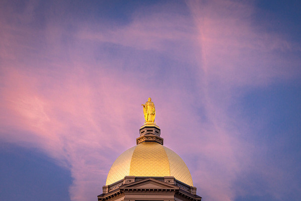 Vivid pink and purple sunset sky behind the Golden Dome of Notre Dame's Main Building with the Virgin Mary statue.