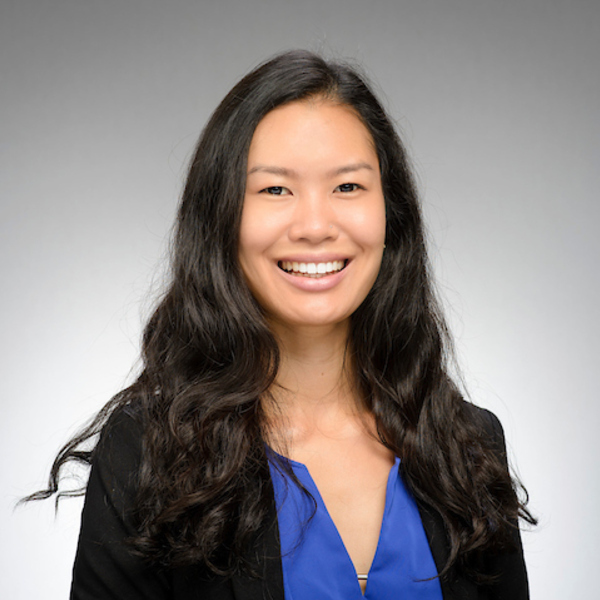 A smiling woman with long dark wavy hair wears a black blazer and a bright blue v-neck top.