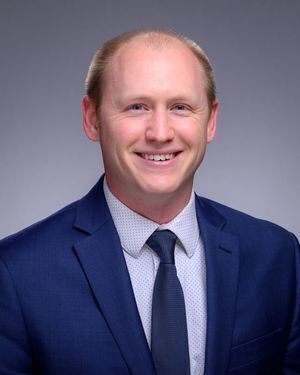Headshot of Caleb Reinking, a smiling man with blonde hair in a blue suit, light blue patterned shirt, and dark blue tie against a gray background.