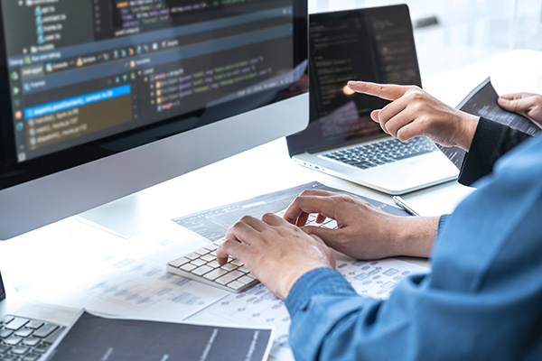 Two people coding. One types on a white keyboard, another points at a laptop screen with code. A large monitor also shows code.