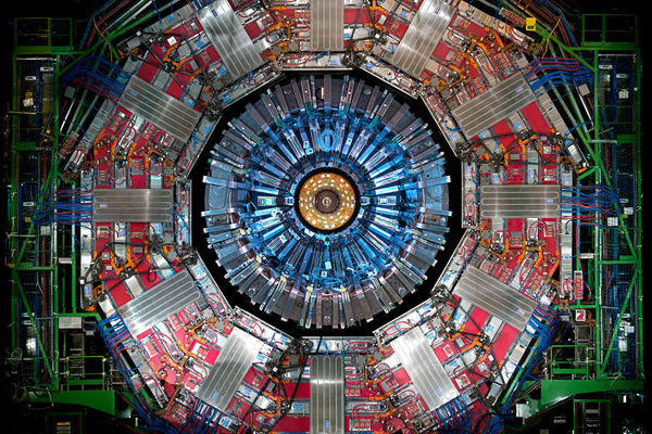 Close-up of a particle detector. Its circular center glows blue, surrounded by complex red, silver, and green machinery.