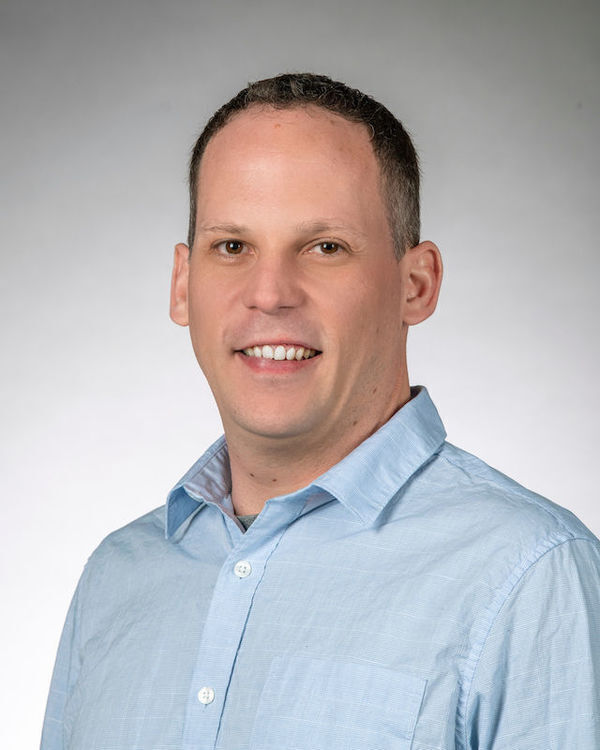 Headshot of Bradley Sandberg, a man with short brown hair, wearing a light blue collared shirt, smiles pleasantly.
