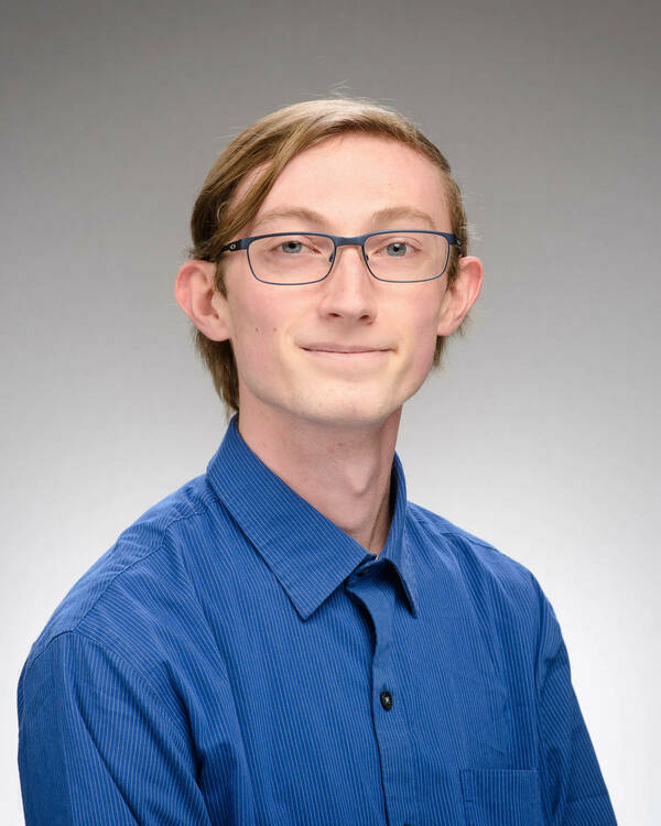 Headshot of Evan Brinckman, a young man with light brown hair, blue glasses, and a blue striped shirt smiles gently.