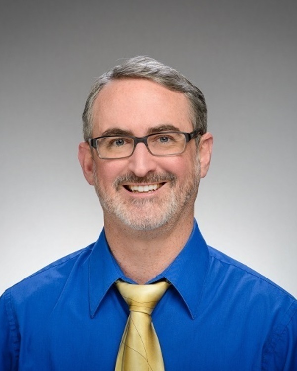Headhsot of Matthew Curran, a man with gray hair and beard, wearing black glasses, a blue shirt, and a gold tie, smiling broadly.