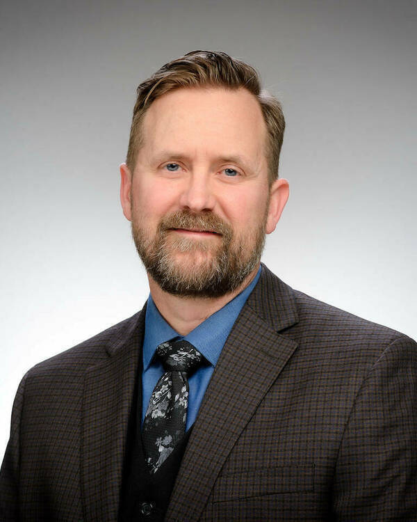 Headhsot of Jared Hendrickson, a bearded man with blue eyes, wearing a blue shirt, black vest, charcoal plaid suit jacket, and floral tie, smiles.