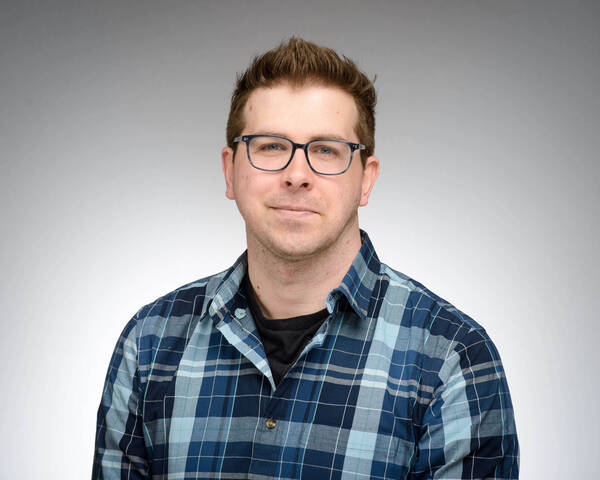 Headshot of Michael Kennel, a young man with brown hair and black glasses, wearing a blue plaid shirt, smiles against a plain gray background.