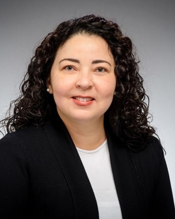Headshot of Priscila Correa Saboia Moreira, a smiling woman with dark curly hair, light skin, and pearl earrings wears a white top and black cardigan.
