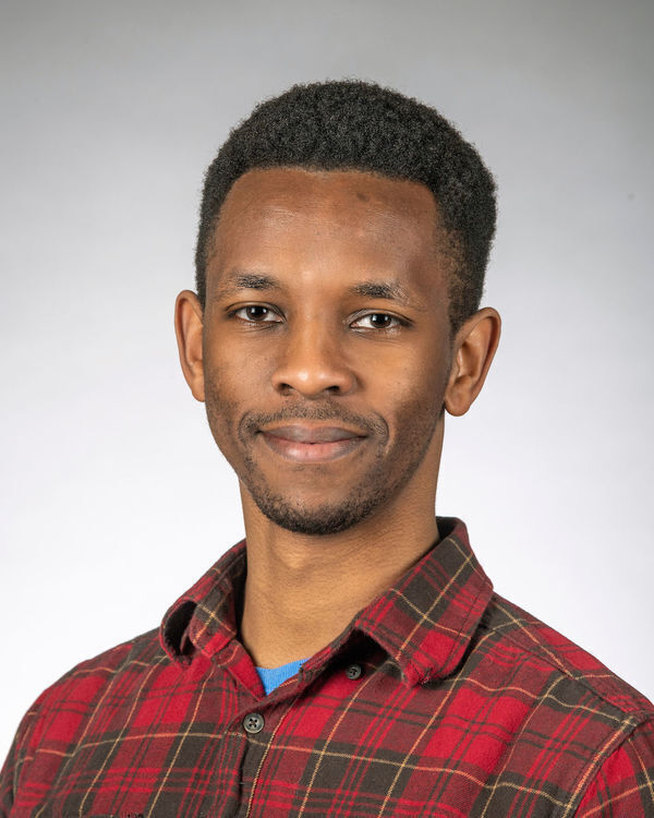 Headshot of Samuel Njoroge, a smiling young Black man with short dark hair and a neatly trimmed beard, wearing a red, brown, and gold plaid shirt.