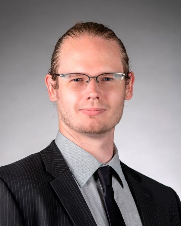 Headshot of James Sweet, a man with light skin, hair in a bun, and glasses wears a pinstripe suit, gray shirt, and dark tie, with a neutral expression.