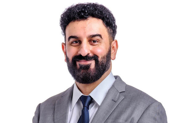 Smiling man with dark curly hair and beard, wearing a gray suit, white shirt, and dark blue tie.