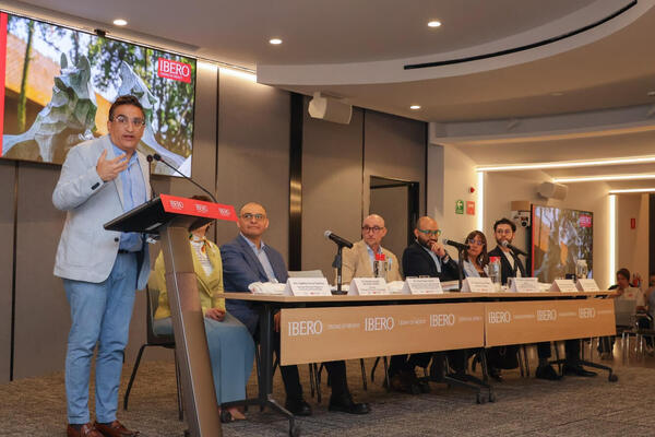 Speaker in light blue blazer gestures at podium. Six panelists sit at a table with IBERO logo, screen displays IBERO Ciudad de México.
