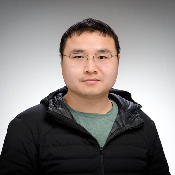 A man with short dark hair and glasses wears a black puffer jacket and a green shirt.
