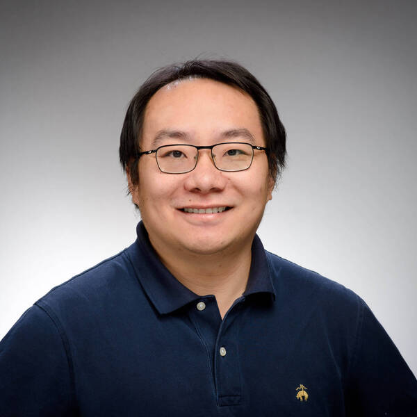 A smiling man with dark hair and glasses wears a navy polo shirt with an embroidered logo against a gray background.