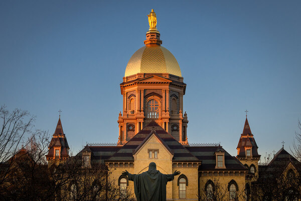 Golden Dome of Notre Dame's Main Building glows in warm light. The Christ the Teacher statue faces the dome, arms outstretched.