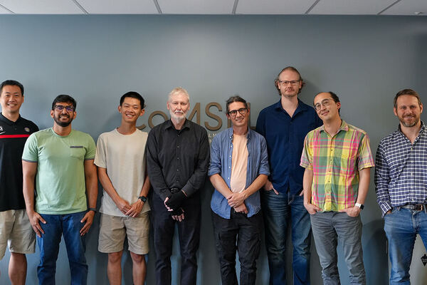 Eight diverse men, some with beards or glasses, stand smiling in casual clothes against a blue-gray wall.