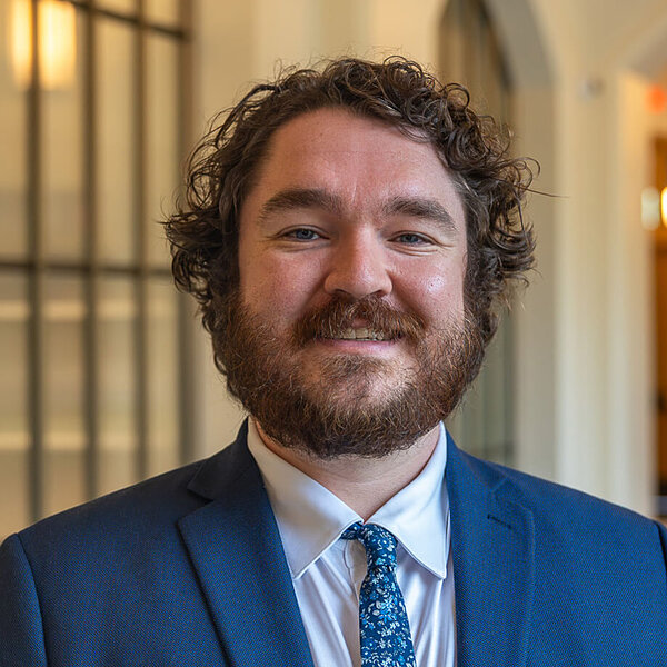 A smiling man with curly brown hair and a dark beard wears a blue suit and floral tie, standing in a building with arched architecture.