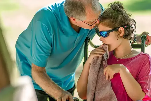 Older man in blue shirt leans toward young person in wheelchair with sunglasses, holding a towel to mouth.