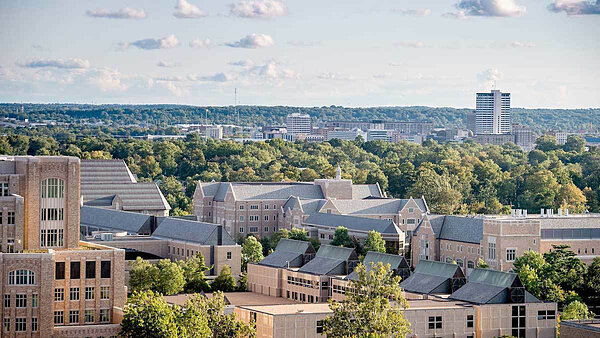 Tops of light brick buildings on the Notre Dame campus with green trees and downtown South Bend in the background.
