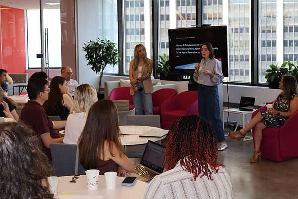 Woman presents to an audience in a modern room with large windows.