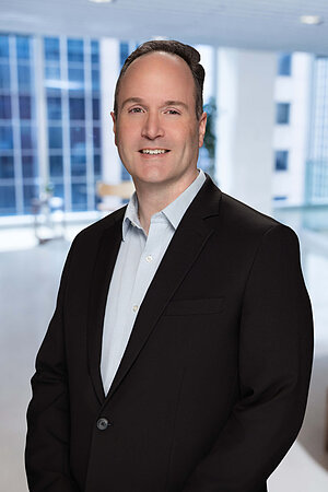 A man in a black suit and blue checkered shirt smiles slightly.