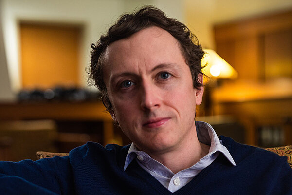 Man with curly brown hair and blue eyes, wearing a dark blue sweater over a light blue striped shirt.